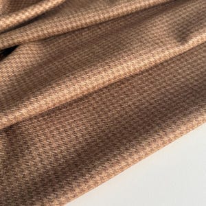 May include: Close-up of folded brown fabric with a houndstooth pattern. The fabric has a textured appearance and a warm color palette, with shades of brown and tan. The material is draped, revealing its texture.