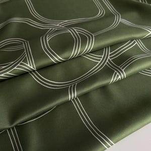 May include: Olive green fabric with a white geometric pattern. The fabric is folded, showing the texture and sheen of the material. The design features interconnected lines and circles.