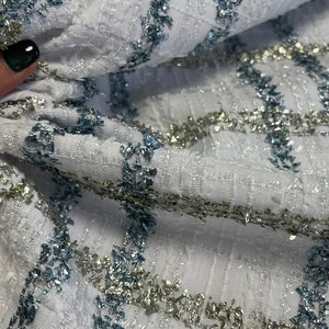 May include: White fabric with a textured weave and silver and blue glitter accents. The fabric has a geometric pattern with stripes of glitter.
