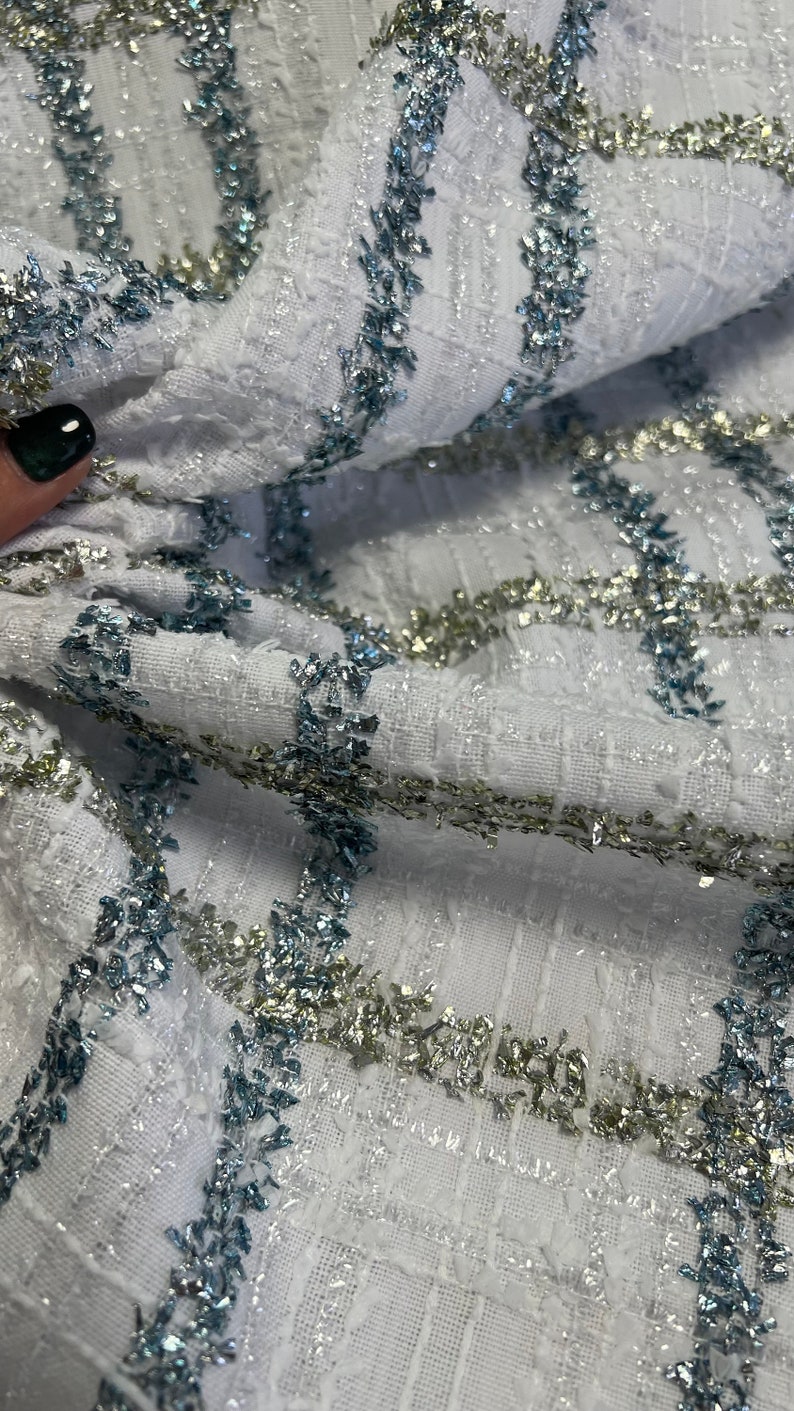 May include: White fabric with a textured weave and silver and blue glitter accents. The fabric has a geometric pattern with stripes of glitter.