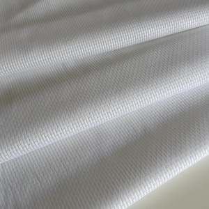 Peut inclure: Gros plan sur un tissu blanc avec un tissage texturé en forme de losanges. Le matériau est plié, montrant la texture et la façon dont la lumière se reflète sur la surface. Le tissu semble doux et léger.