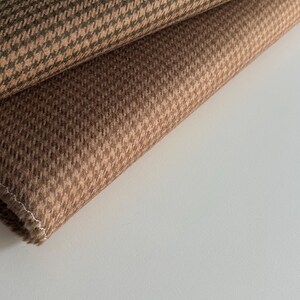 May include: Close-up of two folded pieces of brown houndstooth fabric. The fabric has a repeating pattern of small, broken checks in shades of brown and tan. The fabric is likely used for clothing or upholstery.