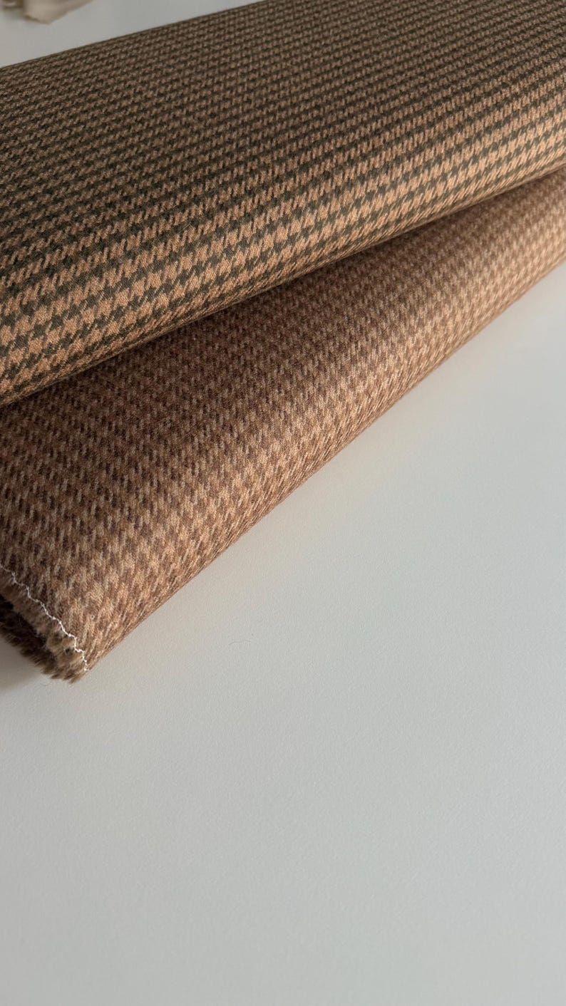 May include: Close-up of two folded pieces of brown houndstooth fabric. The fabric has a repeating pattern of small, broken checks in shades of brown and tan. The fabric is likely used for clothing or upholstery.
