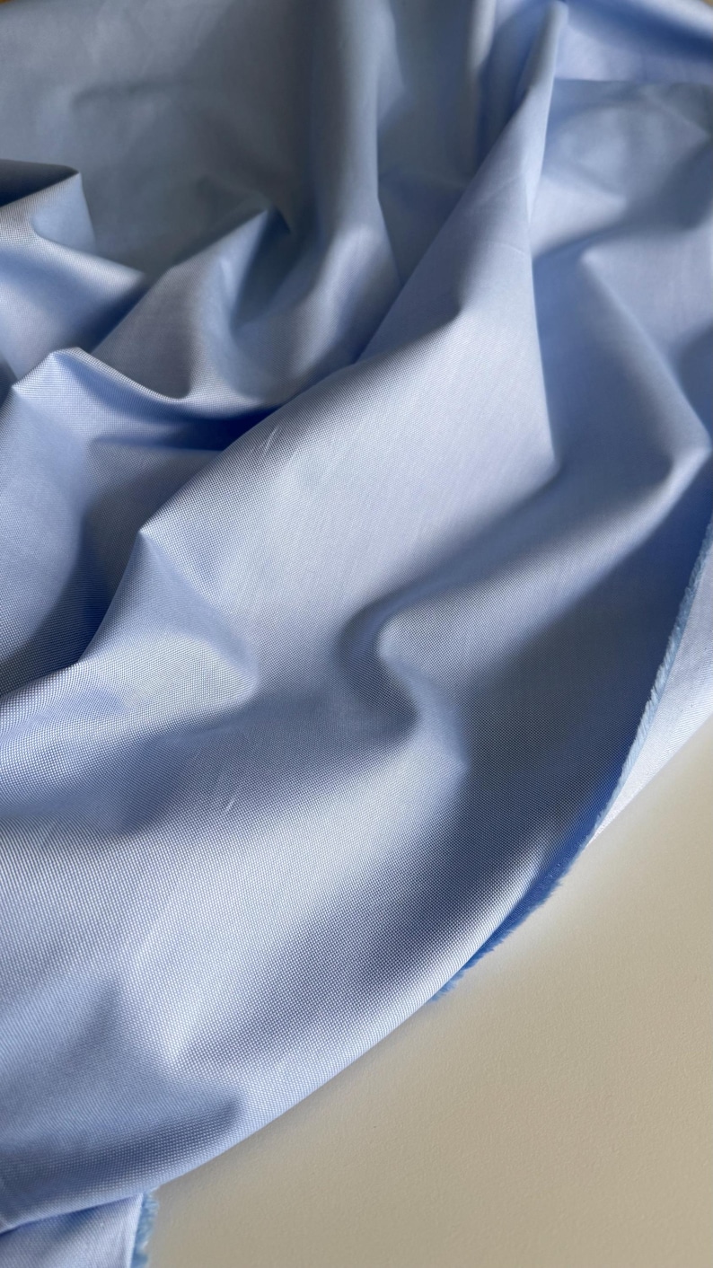 May include: A light blue fabric with a smooth, slightly textured surface. The fabric is folded and draped, showing its soft and flowing nature.