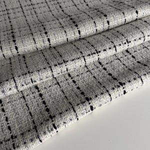 May include: Close-up of folded fabric with a woven texture. The fabric is mainly light grey with a grid pattern of black lines. The fabric is layered, showing the texture and weave of the material.