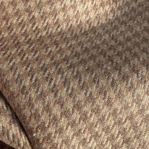 May include: Close-up of a textured fabric with a herringbone pattern. The fabric is a blend of light and dark brown tones, creating a subtle contrast. The weave is visible, adding depth and dimension to the material.