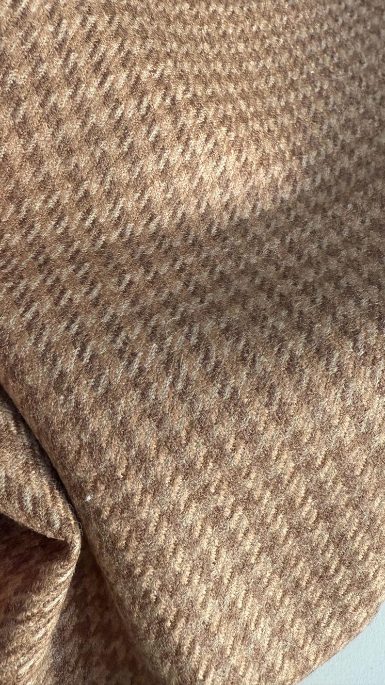 May include: Close-up of a textured fabric with a herringbone pattern. The fabric is a blend of light and dark brown tones, creating a subtle contrast. The weave is visible, adding depth and dimension to the material.