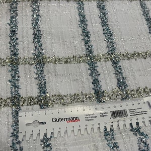 May include: White fabric with a grid pattern, accented with silver and blue glitter. The fabric is measured with a ruler in inches.