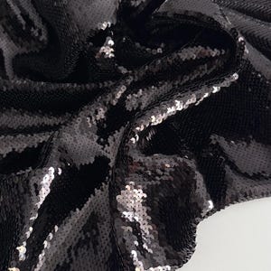 Peut inclure: Gros plan sur un tissu à paillettes noir. Le tissu est drapé, montrant la texture et la qualité réfléchissante des paillettes. Les paillettes sont densément tassées, créant un effet scintillant. Le fond est une surface blanche unie.