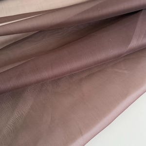 Könnte beinhalten: Nahaufnahme von mehrlagigem, transparentem Stoff in Braun- und Mauve-Tönen. Der Stoff ist gefaltet und drapiert, wodurch weiche Schatten und Highlights entstehen. Das Material wirkt leicht und zart, geeignet für Bekleidung oder Dekorationsprojekte.
