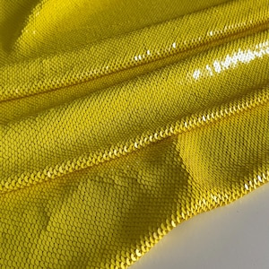 Può includere: Un primo piano di un tessuto giallo con una texture squamosa. Il tessuto è fatto di paillettes ed è piegato a metà.