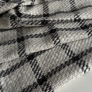 Op de afbeelding: Close-up van een textiel met een ruitpatroon. De stof is voornamelijk lichtgrijs met zwarte en zilveren accenten. De weefsel is zichtbaar en de stof is gevouwen, wat diepte en textuur laat zien. De algehele uitstraling is elegant en modern.