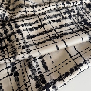 Könnte beinhalten: Nahaufnahme eines cremefarbenen Stoffes mit einem schwarzen, pinselstrichartigen Gittermuster. Der Stoff ist gefaltet und zeigt seine Textur und seinen Glanz. Das abstrakte, moderne Design eignet sich für Kleidung oder Heimtextilien.