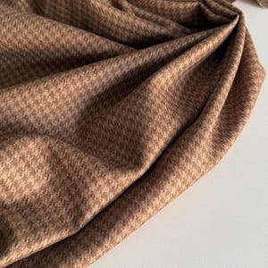 May include: Close-up of a folded brown fabric with a subtle houndstooth pattern. The fabric is draped, creating soft folds and shadows. The color is a warm, neutral brown, suitable for various sewing projects.