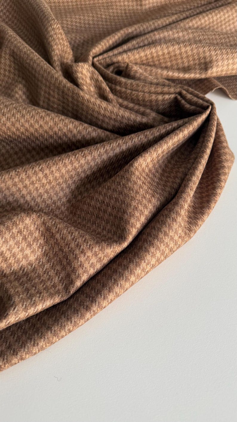 May include: Close-up of a folded brown fabric with a subtle houndstooth pattern. The fabric is draped, creating soft folds and shadows. The color is a warm, neutral brown, suitable for various sewing projects.