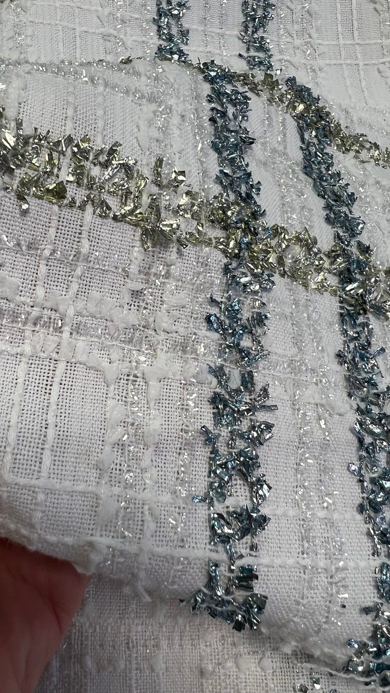 May include: White fabric with a woven texture and silver and blue embellishments. The fabric has a subtle checkered pattern.