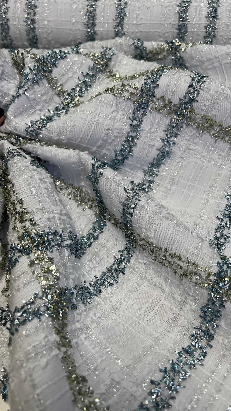 May include: White fabric with a textured weave and silver and blue glitter stripes. The fabric is folded and the glitter stripes are visible.