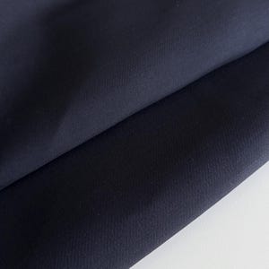 Może przedstawiać: Zbliżenie granatowej tkaniny, złożonej, aby ukazać fakturę i splot. Tkanina ma głęboki, jednolity kolor z subtelnymi odcieniami. Zagięcia tworzą cienie i refleksy, podkreślając układ i ciężar materiału.