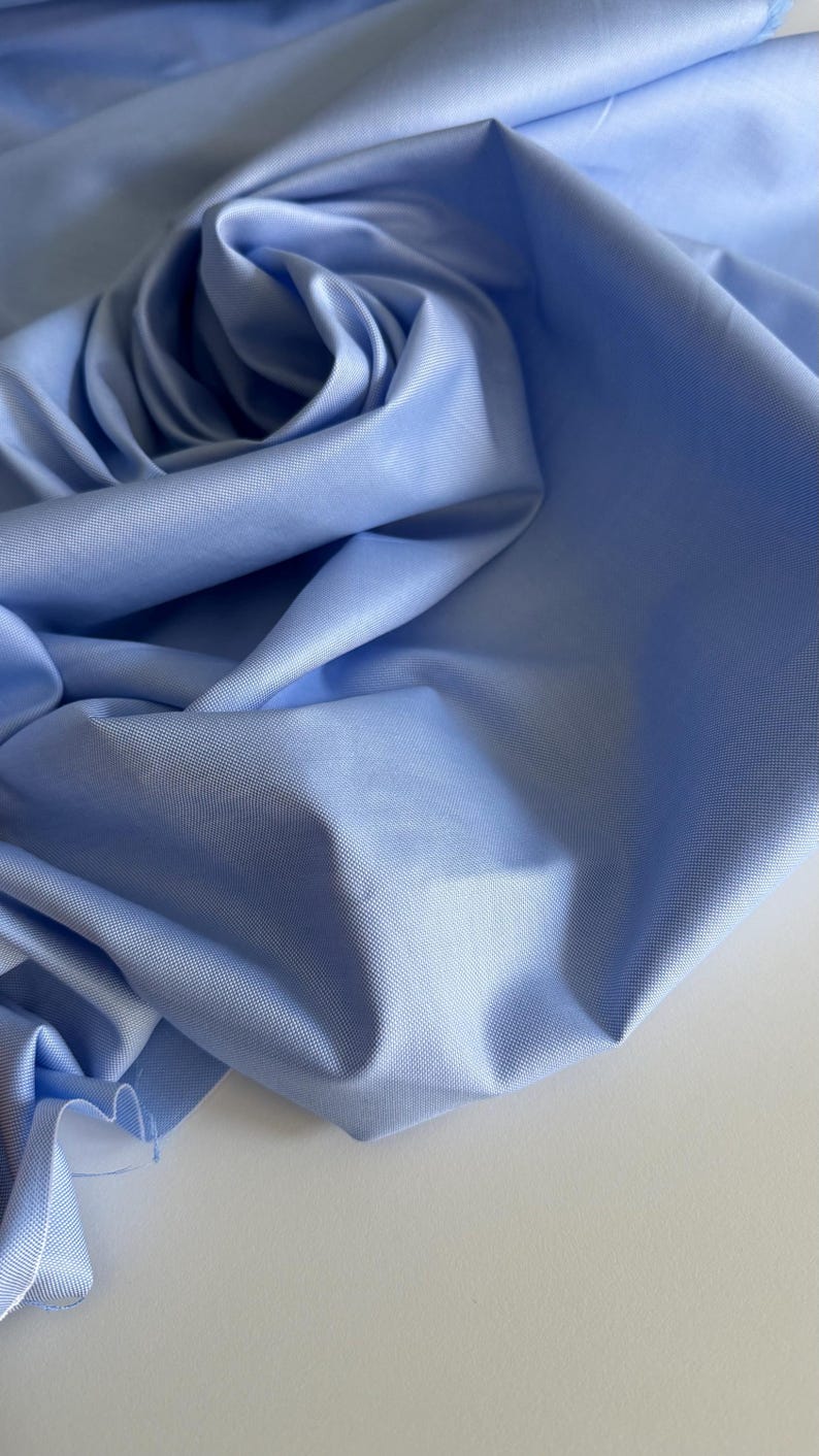 May include: A close-up of a light blue fabric with a smooth, textured surface. The fabric is folded and draped, creating a soft, flowing look.