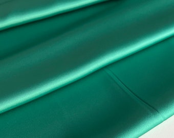 May include: A smooth, folded piece of teal satin fabric. The fabric has a subtle sheen and is draped over a white surface.