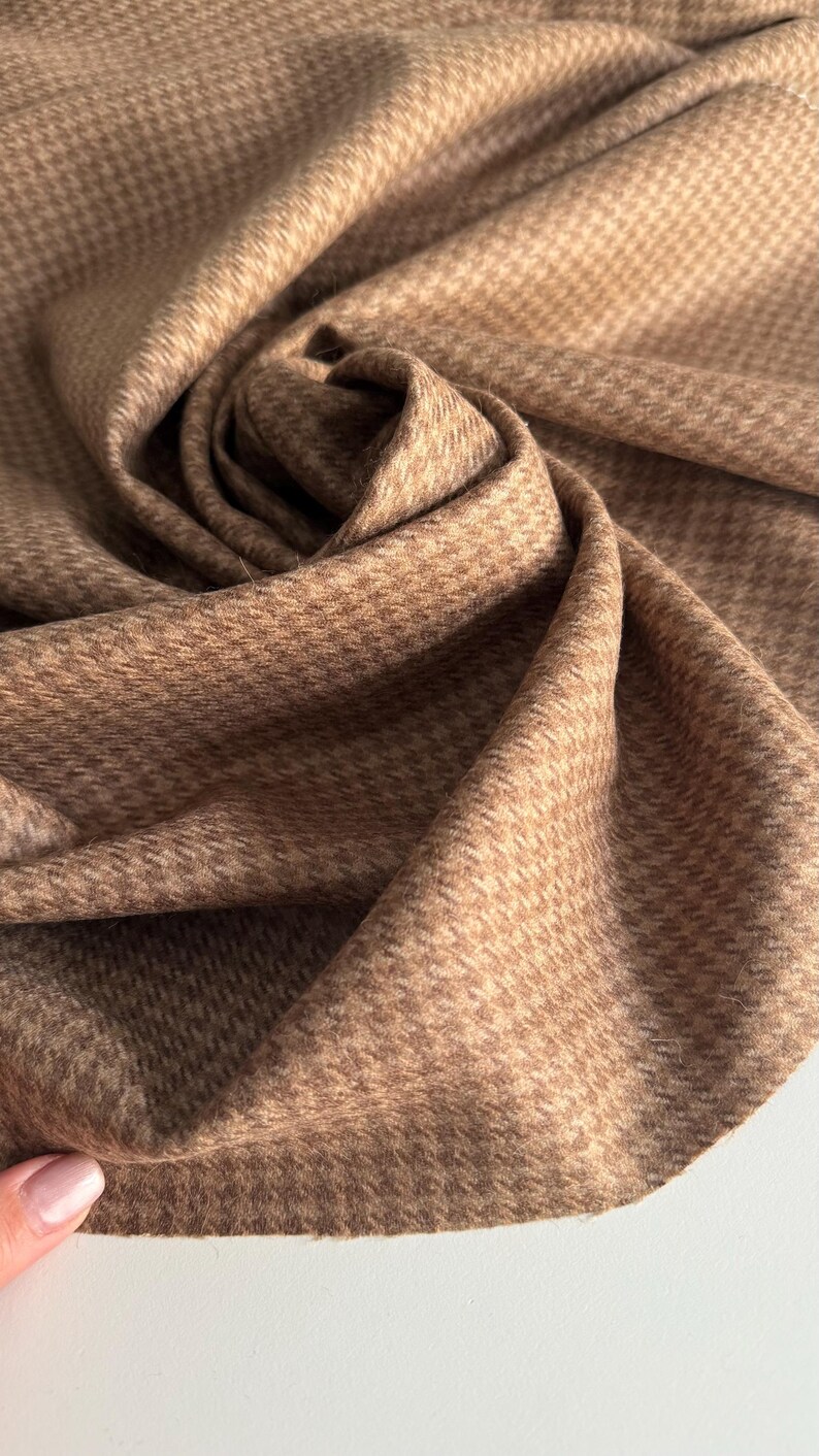 May include: Close-up of a folded fabric with a houndstooth pattern in shades of brown and tan. The fabric is soft and textured, with a subtle sheen. The pattern is small and evenly spaced, creating a classic and elegant look. Ideal for apparel and home decor.