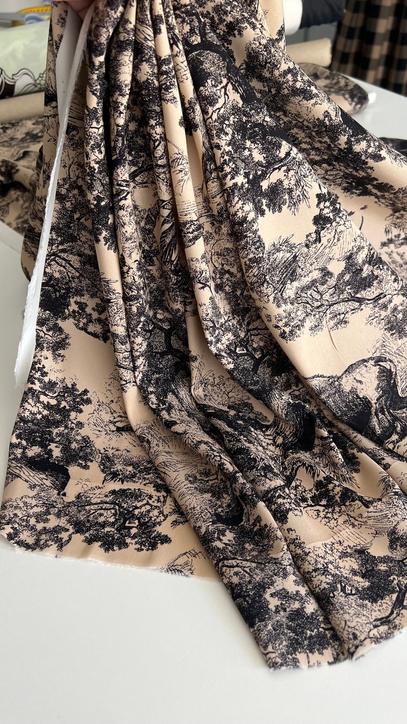 May include: Beige fabric with a black and white print of trees and foliage. The fabric is folded and draped on a white surface.
