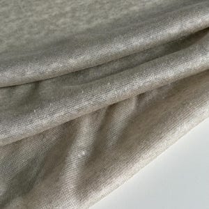Könnte beinhalten: Nahaufnahme eines gefalteten, hellbeigen Stoffes. Das Material wirkt weich und leicht strukturiert, mit subtilen Farbvariationen. Der Stoff ist drapiert, wodurch Falten und Schatten entstehen, die dem Bild Tiefe verleihen. Der Gesamteindruck ist neutral und vielseitig.