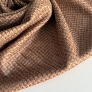 May include: Close-up of a folded fabric with a houndstooth pattern in shades of brown and beige. The fabric is soft and appears to be draped, with visible folds and textures. The background is a plain white surface.