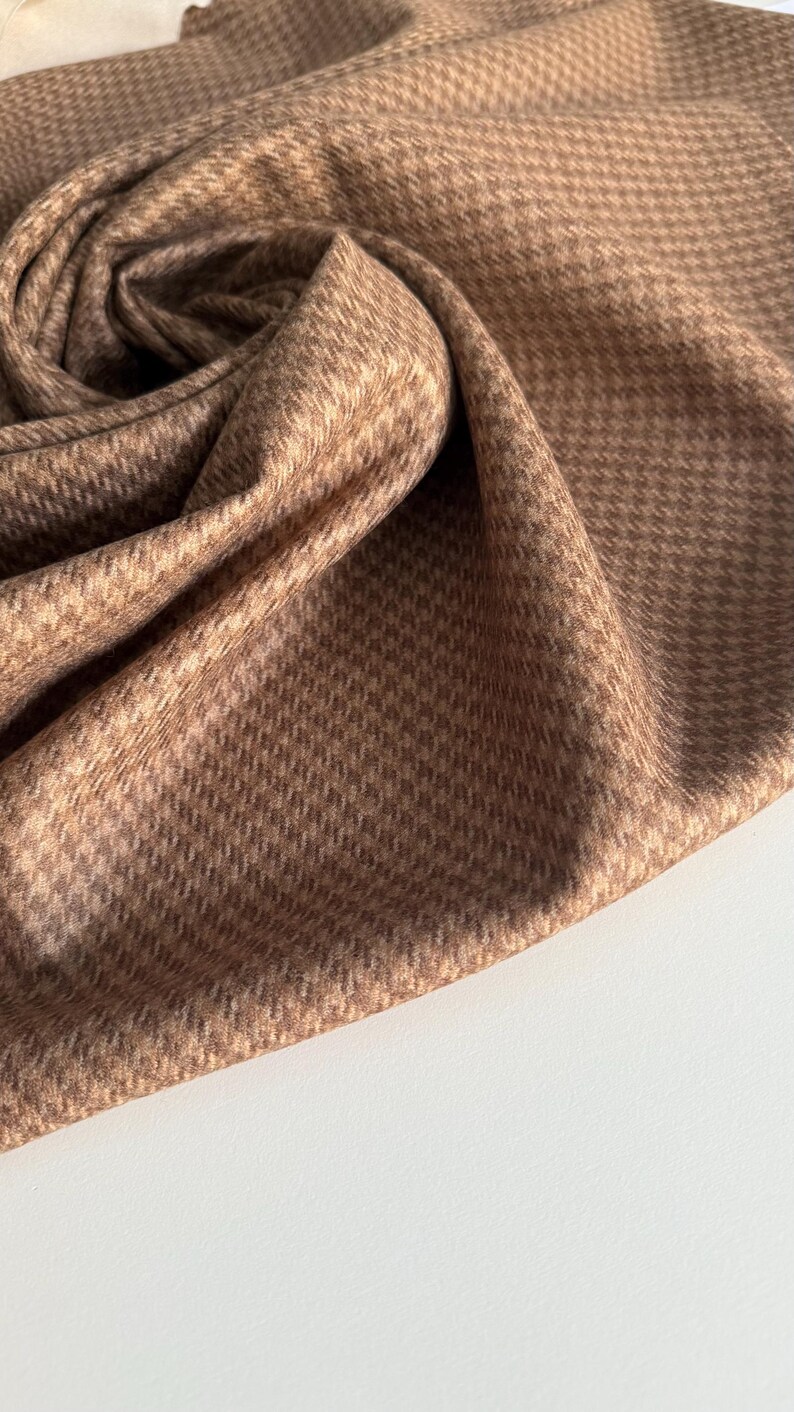 May include: Close-up of a folded fabric with a houndstooth pattern in shades of brown and beige. The fabric is soft and appears to be draped, with visible folds and textures. The background is a plain white surface.