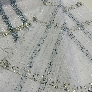 May include: White fabric with a silver and blue checkered pattern. The fabric has a textured surface and a slightly frayed edge.