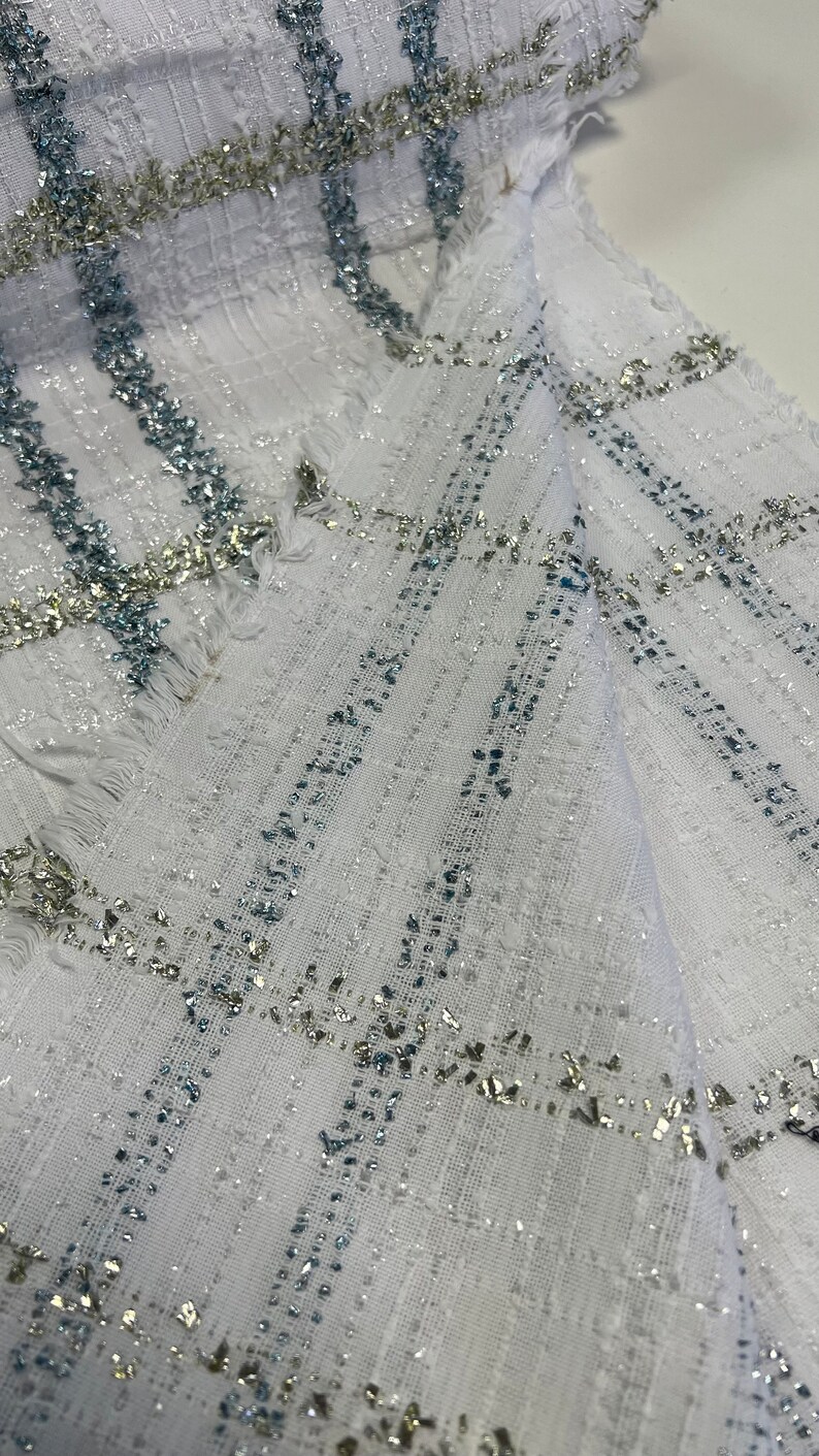 May include: White fabric with a silver and blue checkered pattern. The fabric has a textured surface and a slightly frayed edge.