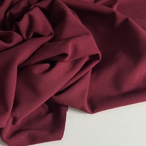 Pu&ograve; includere: Primo piano di un tessuto color borgogna scuro con superficie testurizzata. Il tessuto &egrave; piegato e drappeggiato su una superficie bianca.