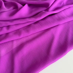Könnte beinhalten: Ein leuchtend pinkfarbener Stoff mit weichen Falten und dezentem Glanz. Das Material wirkt leicht und fließend, was auf eine Verwendung für Kleidung oder Dekorationszwecke hindeutet. Die Stoffstruktur ist glatt, mit sanften Schatten, die Tiefe erzeugen.