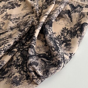 May include: Beige fabric with a black and white print of a forest scene. The fabric is folded and draped on a white surface.
