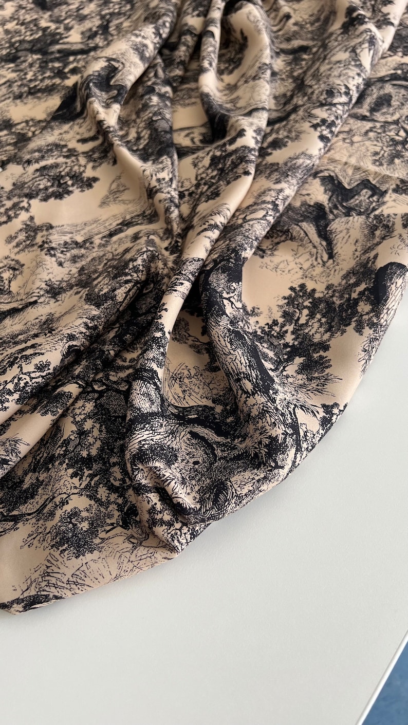 May include: Beige fabric with a black and white print of a forest scene. The fabric is folded and draped on a white surface.