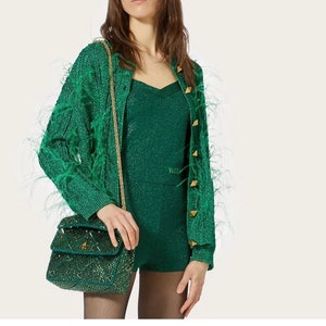 May include: A green feather-trimmed cardigan with gold buttons and a matching green sequined mini dress. The model is carrying a small green sequined handbag with a gold chain strap.