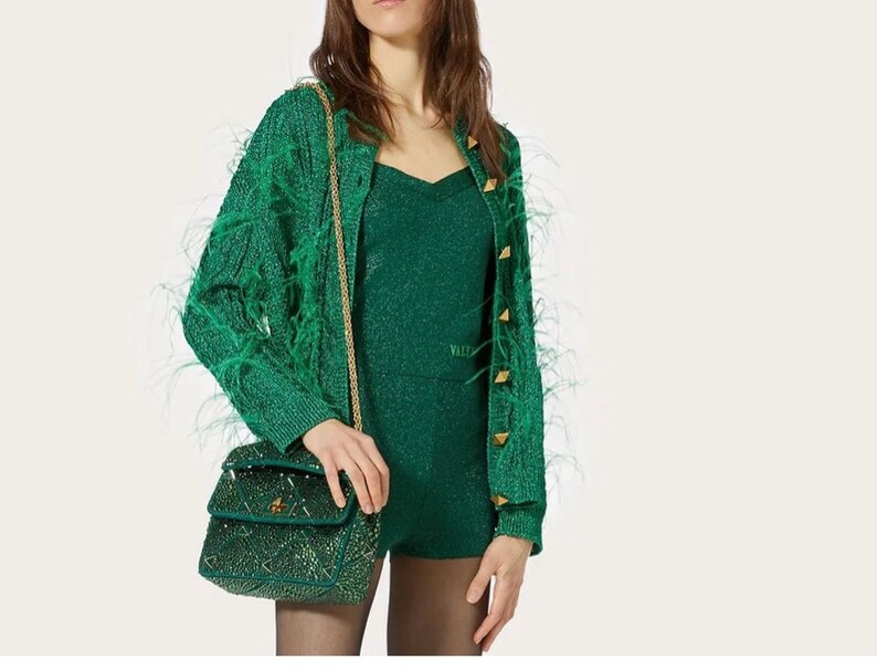 May include: A green feather-trimmed cardigan with gold buttons and a matching green sequined mini dress. The model is carrying a small green sequined handbag with a gold chain strap.