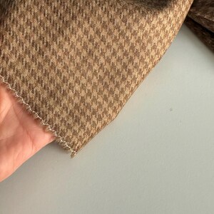 May include: Close-up of a brown and tan houndstooth fabric. The fabric is folded, revealing a detailed pattern. The edges are finished with a white, zig-zag stitch. The fabric has a soft, textured appearance.