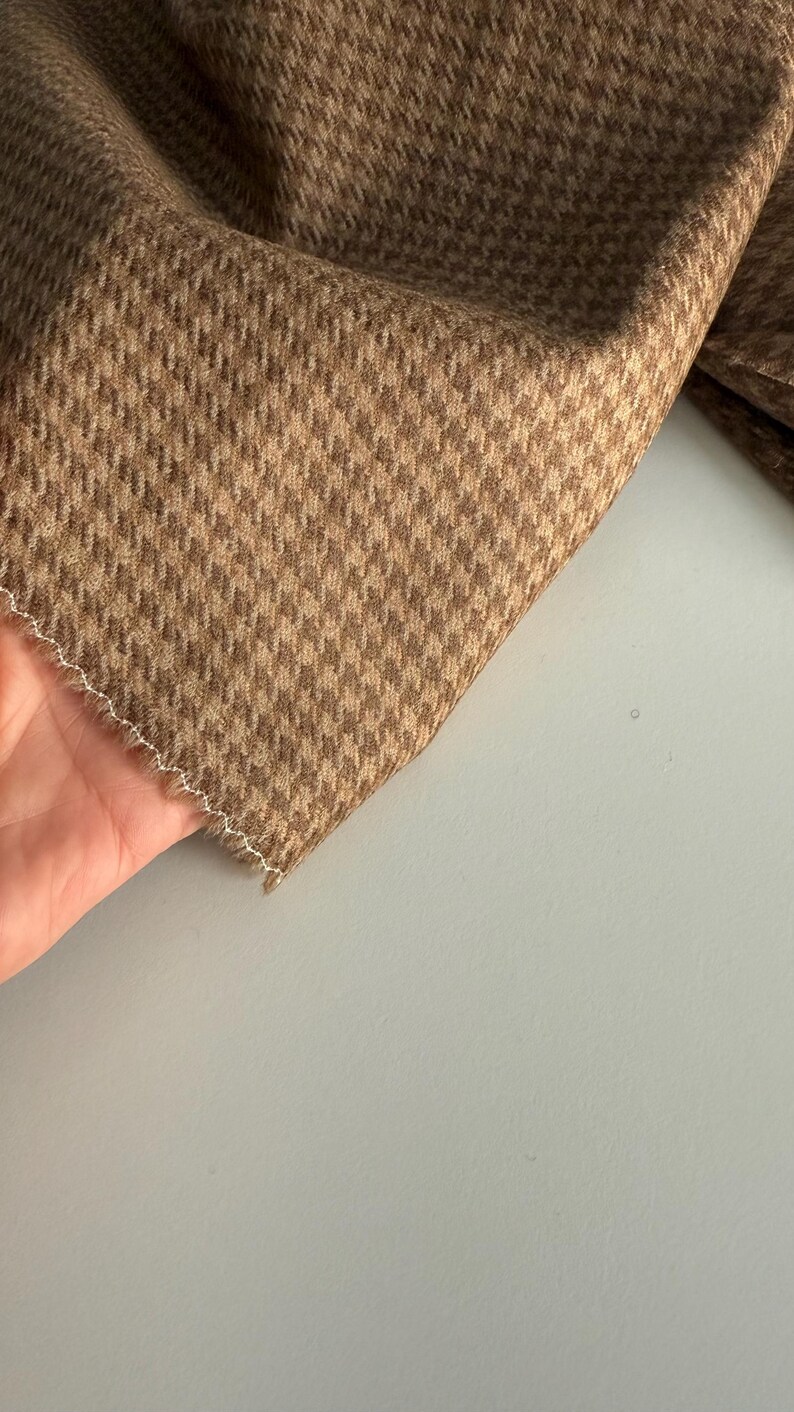 May include: Close-up of a brown and tan houndstooth fabric. The fabric is folded, revealing a detailed pattern. The edges are finished with a white, zig-zag stitch. The fabric has a soft, textured appearance.