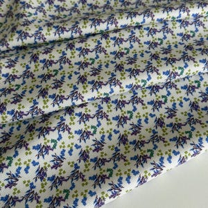 Könnte beinhalten: Nahaufnahme eines weißen Stoffes mit einem sich wiederholenden Blumenmuster. Das Design zeigt kleine blaue, violette und grüne Blumen und Blätter, die über die Oberfläche verstreut sind. Der Stoff ist gefaltet und zeigt Textur und Tiefe.