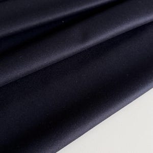 Könnte beinhalten: Nahaufnahme eines dunkelmarineblauen Stoffes mit weichen Falten. Das Material erscheint glatt und undurchsichtig, mit subtilen Farbtonvariationen. Der Stoff wird wahrscheinlich für Kleidung oder Heimdekoration verwendet.
