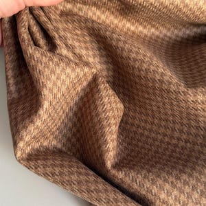 May include: Close-up of a folded brown fabric with a houndstooth pattern. The fabric is a warm, earthy tone, with a subtle texture. The pattern is a classic, small-scale design, creating a sophisticated look. The fabric appears soft and pliable.