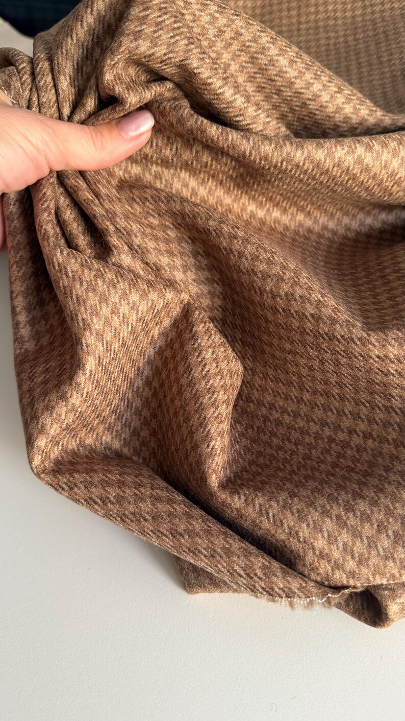 May include: Close-up of a folded brown fabric with a houndstooth pattern. The fabric is a warm, earthy tone, with a subtle texture. The pattern is a classic, small-scale design, creating a sophisticated look. The fabric appears soft and pliable.