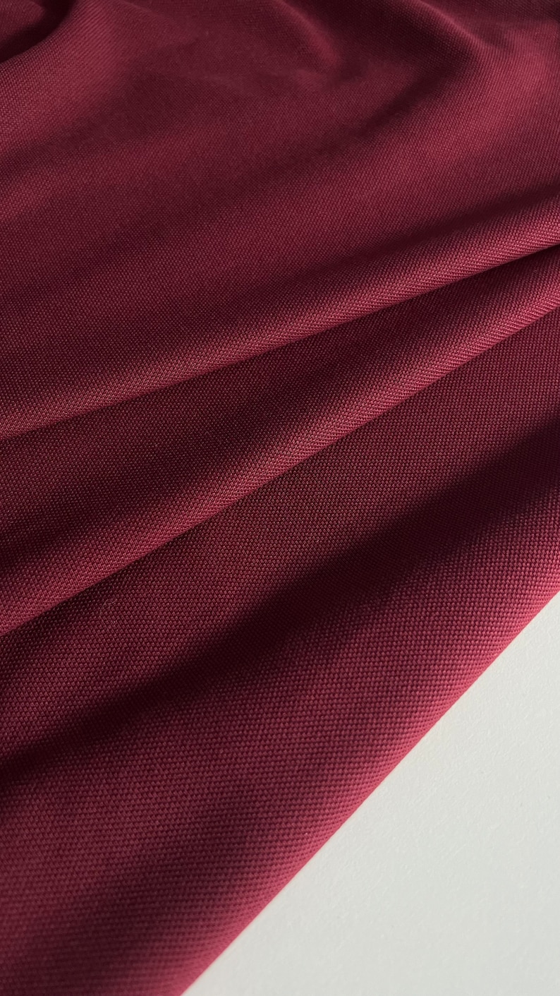 Pu&ograve; includere: Primo piano di un tessuto color borgogna scuro con una trama testurizzata. Il tessuto &egrave; piegato e drappeggiato, mostrando la sua texture morbida e flessibile.
