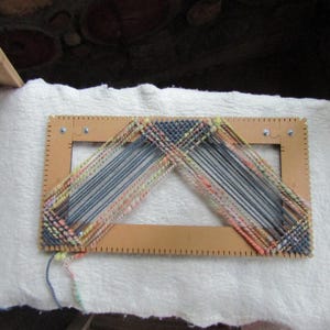 May include: A rectangular wooden weaving loom frame with notched edges, featuring a partially completed woven project made of multi-colored yarn in shades of blue, pink, yellow, and green, arranged in a triangular pattern across the center opening.