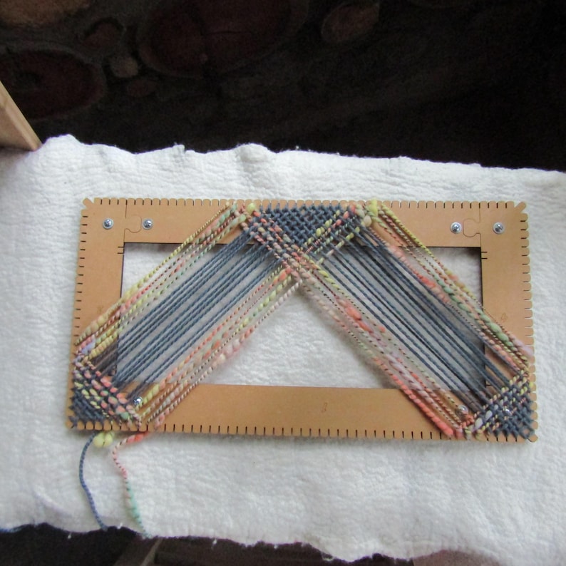 May include: A rectangular wooden weaving loom frame with notched edges, featuring a partially completed woven project made of multi-colored yarn in shades of blue, pink, yellow, and green, arranged in a triangular pattern across the center opening.
