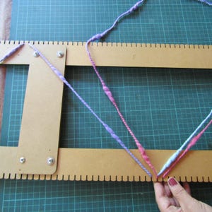 May include: A tan wooden weaving loom with evenly spaced notches along the edges, featuring a length of variegated yarn in shades of purple, pink, and light blue threaded through the notches in a V-shaped pattern, secured with metal screws.