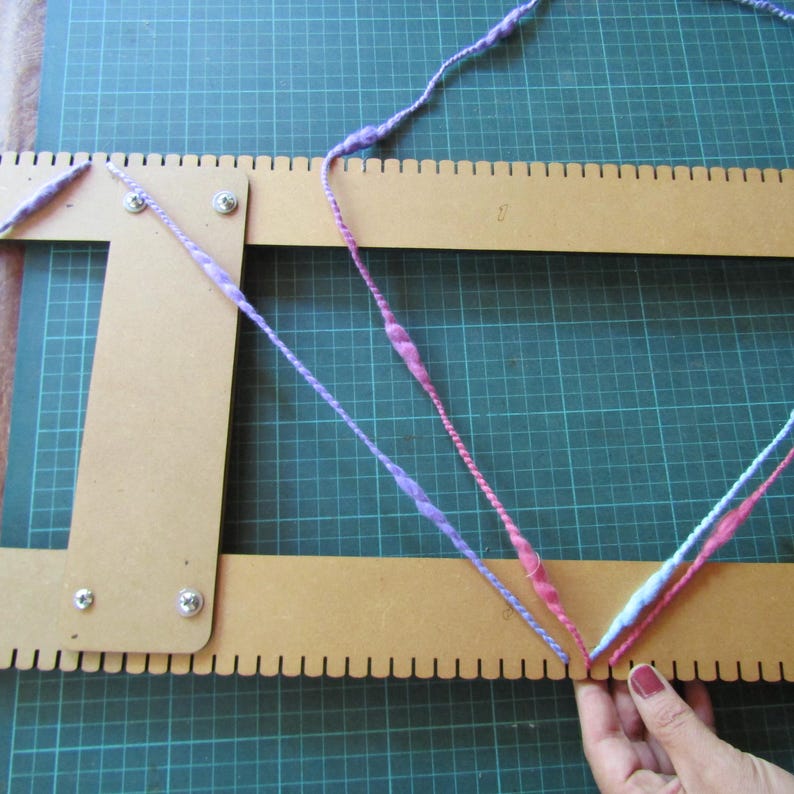 May include: A tan wooden weaving loom with evenly spaced notches along the edges, featuring a length of variegated yarn in shades of purple, pink, and light blue threaded through the notches in a V-shaped pattern, secured with metal screws.