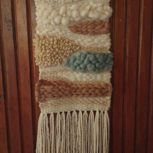 May include: A hand-woven wall hanging featuring a variety of textures and yarn weights in cream, beige, dusty rose, and muted blue. The piece is suspended from a wooden dowel with small wooden beads and finished with long cream fringe at the bottom.