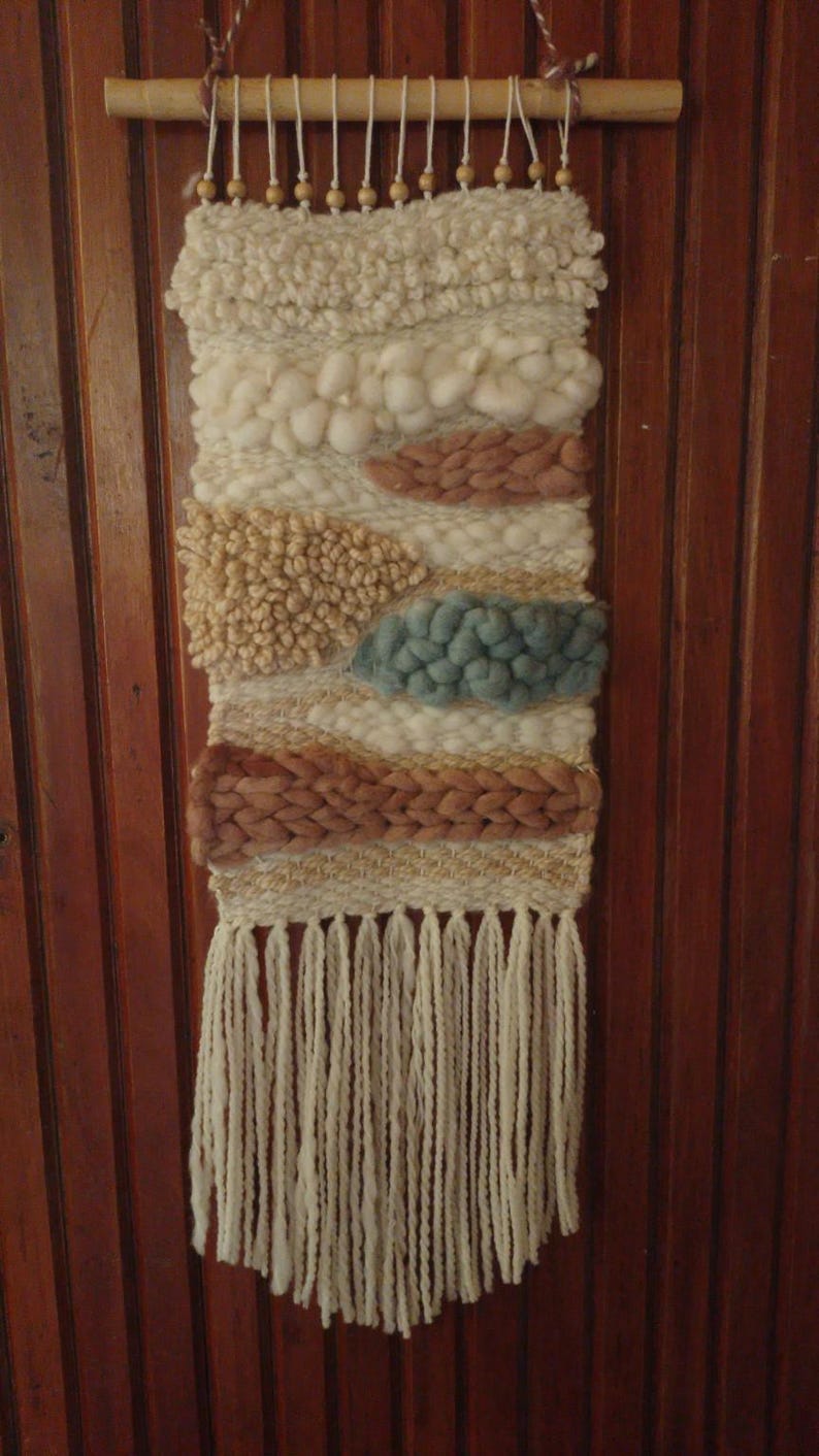 May include: A hand-woven wall hanging featuring a variety of textures and yarn weights in cream, beige, dusty rose, and muted blue. The piece is suspended from a wooden dowel with small wooden beads and finished with long cream fringe at the bottom.